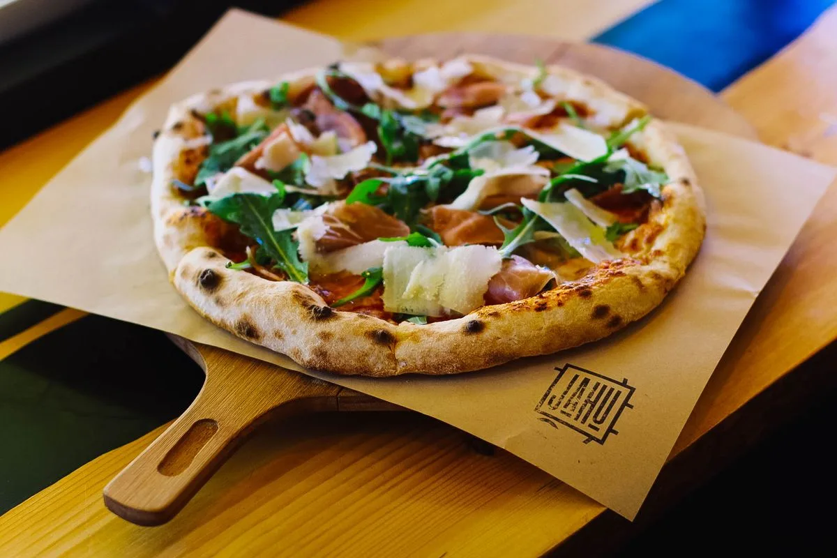 Pizza artesanal recién horneada sobre una pala de madera con jamón y rúcula